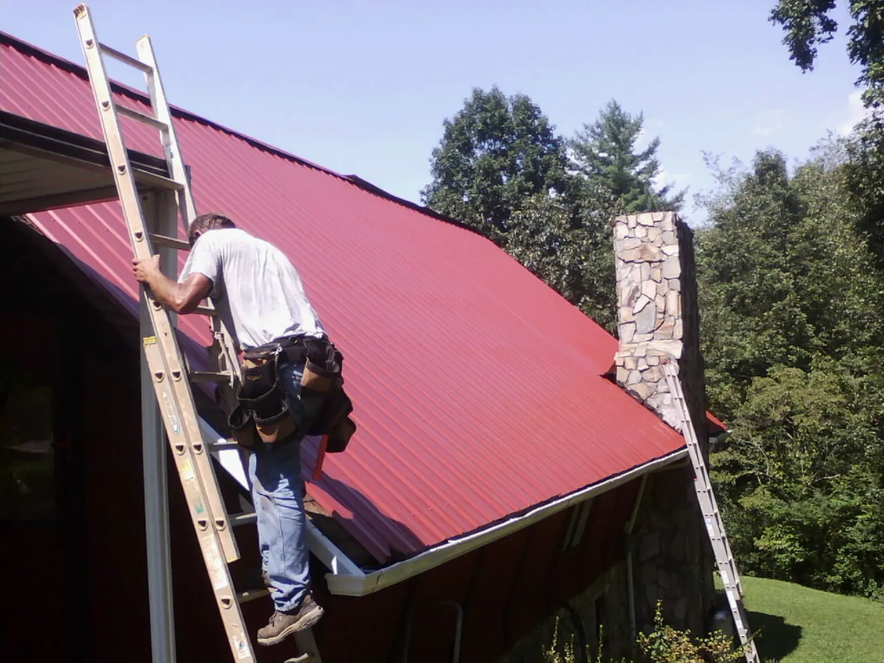 Roofer on a ladder beside a red metal roof and stone chimney — Soffit & Fascia Repair by Roofing Brothers in Wadsworth